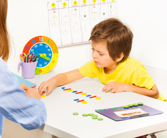 Concentrated boy putting colorful coins in order during developing game with his mother sitting at the table indoors